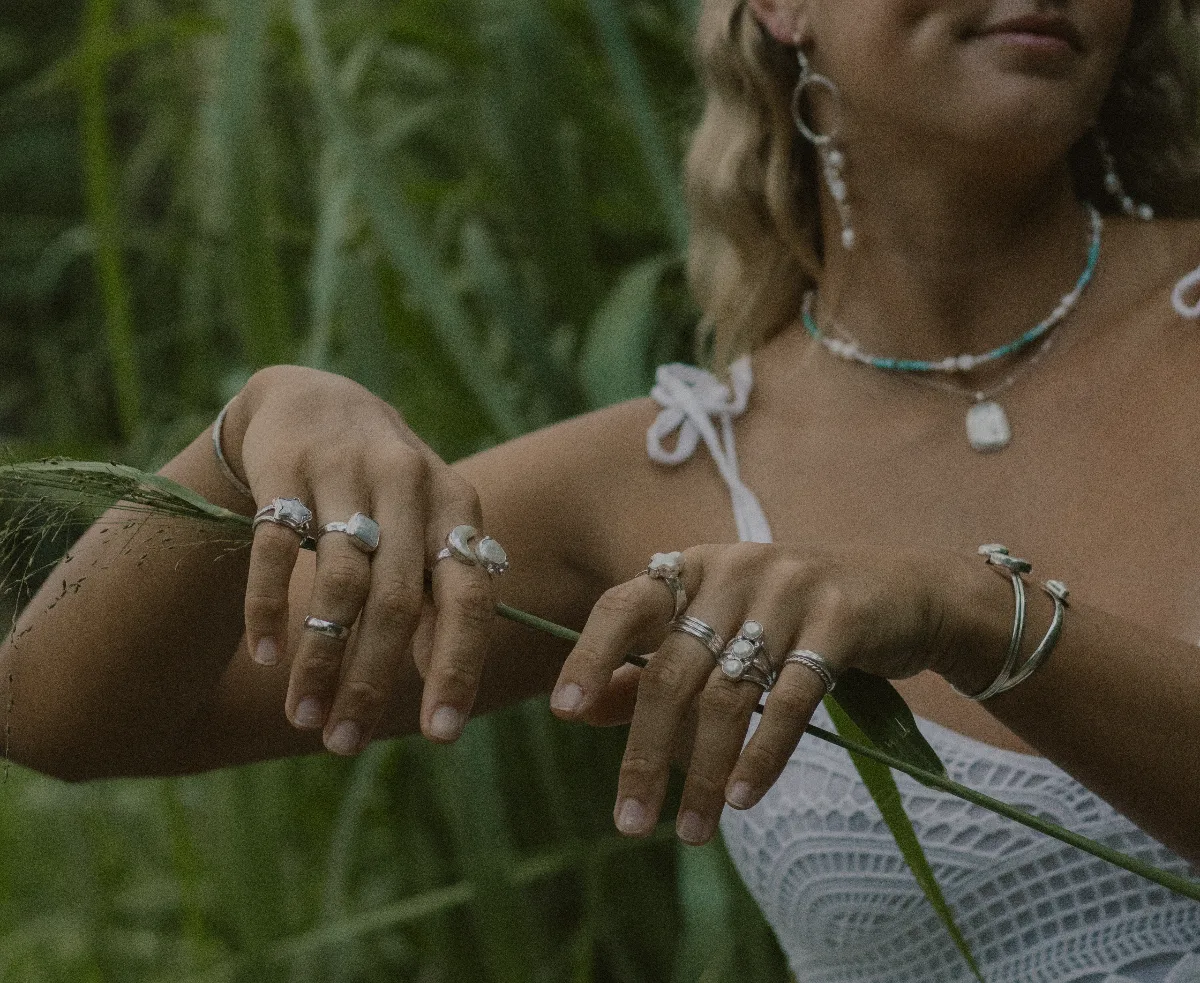 A woman crouched at the water's edge on a jungle-lined beach, wearing silver rings and cuffs